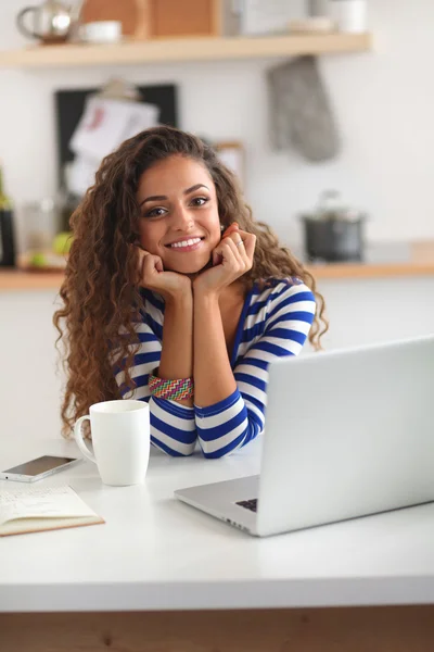 Smiling young woman with coffee cup and laptop in the kitchen at home — Stock Photo, Image