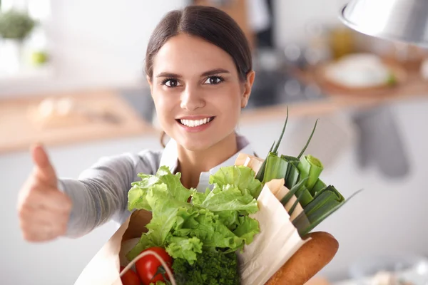 Mujer joven sosteniendo bolsa de la compra de comestibles con verduras. De pie en la cocina — Foto de Stock