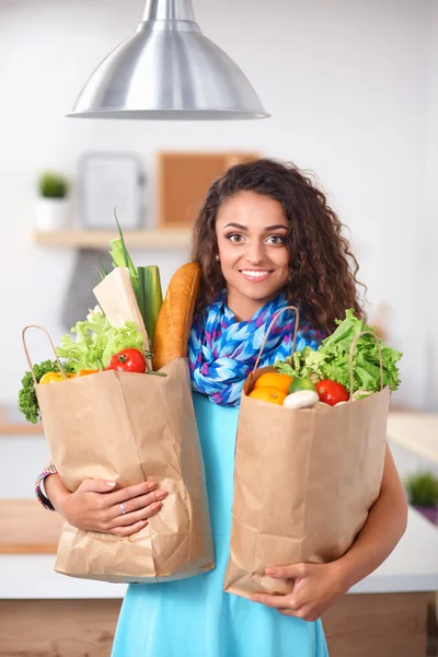 Ung kvinde holder købmand indkøbspose med grøntsager. Stående i køkkenet - Stock-foto