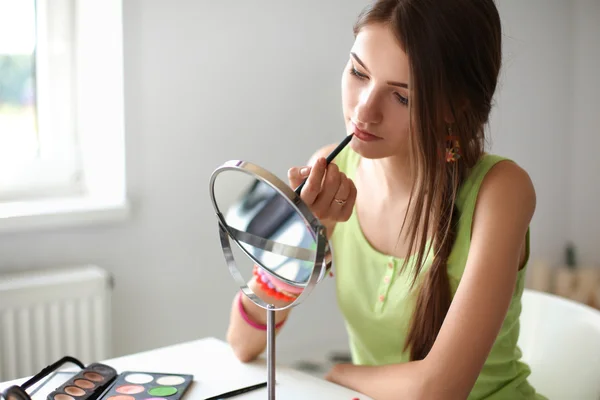 Ung smuk kvinde gør make-up nær spejl, sidder ved skrivebordet - Stock-foto