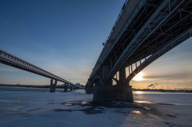 Şehrin arka planına karşı çarpıcı bir gün batımı. Arka planda bir demiryolu köprüsü, nehirde yüzen gemiler ve çok katlı binalar görebilirsiniz.. 