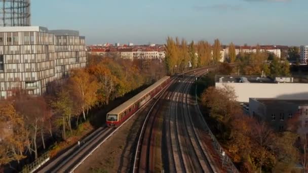 旅客列車は、オフィスビルの隣に公共交通機関、空中ドリーでゴールデンアワーの夕日でドイツのビッグシティベルリンの木によって通過落書き列車 — ストック動画