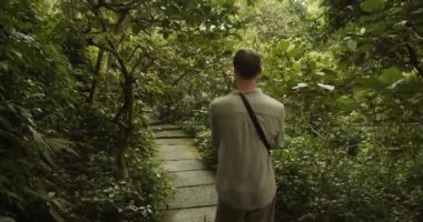 Tropikal yağmur ormanlarında taş yolda yürüyen genç bir adamın elde çekilen görüntüsünü takip ediyoruz.