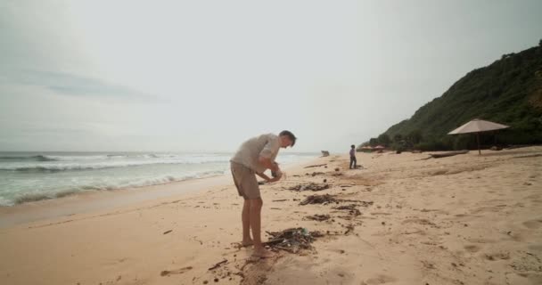 Vista de cerca de un joven caucásico recogiendo basura en la playa. Hombre limpieza turística contaminación plástica en la playa de Bali — Vídeo de stock