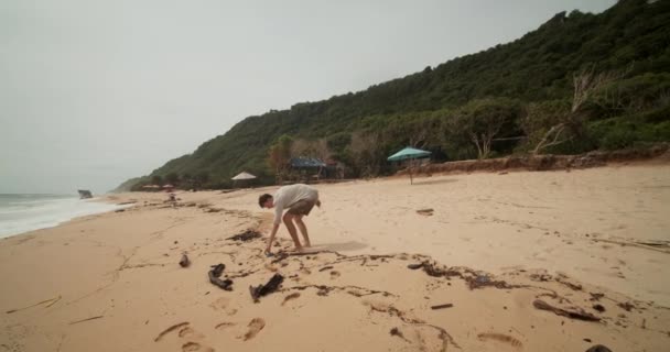 Homem branco jovem pegando o lixo em uma praia de areia em Bali. A aproximar-se de um homem de camisa cáqui e calções a limpar a praia — Vídeo de Stock