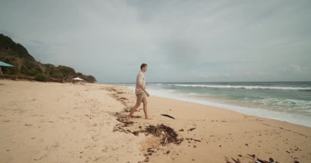Jovem macho branco em shorts cáqui e camisa caminhando em direção ao oceano na bela praia tropical em Bali com algas — Vídeo de Stock
