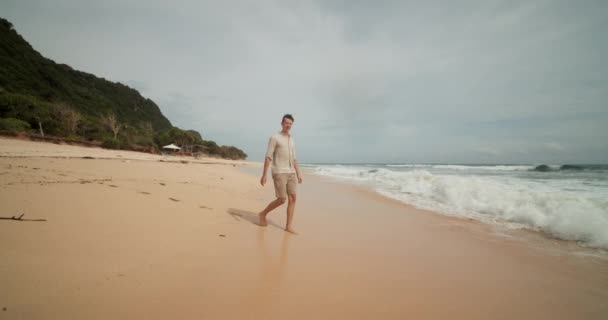 Jonge aantrekkelijke man wandelen op het natte zand in de oceaan deining in het strand in Uluwatu, Bali en stoppen in de golven — Stockvideo