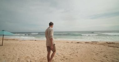 Genç beyaz erkek Bali 'de kumsalda haki şort ve tişörtle yürüyor.