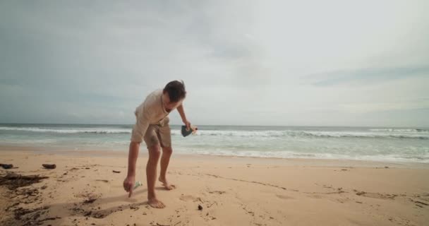 Közelkép egy férfit követve, aki feltakarítja a partot és felszedi a törmeléket a Bali homokos partján lévő üledékekből. — Stock videók