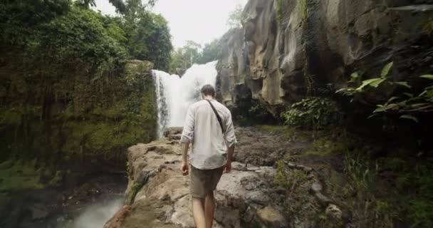 Seguimiento de disparo de mano siguiendo a un joven caminando sobre el acantilado de piedra hacia la cascada en una selva tropical. — Vídeos de Stock
