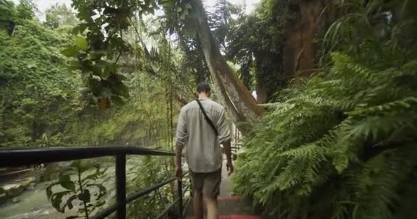 Nach Handschüssen auf einen Mann, der auf der Steintreppe in einem tropischen Regenwald mit einem großen Wasserfall im Vordergrund geht — Stockvideo