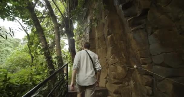 Nach einem Schuss auf den Rücken eines jungen Mannes, der auf einer Treppe entlang einer Steinklippe in einem tropischen Regenwald auf Bali, Indonesien, spaziert — Stockvideo