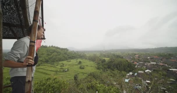 Homem desfrutando da vista do topo da cabana de observação de madeira acima do deslumbrante campo de Bali com plantações de fazenda de arroz e bairros residenciais — Vídeo de Stock