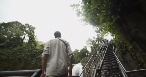 Basso angolo di scatto di un giovane in piedi su una scala di metallo con la vista della grande cascata nella giungla — Video Stock