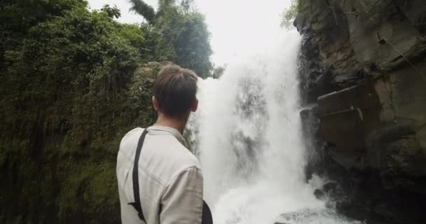 Sudut rendah ditembak berputar-putar di sekitar seorang penjelajah laki-laki muda menunjuk pada air terjun besar di hutan — Stok Video