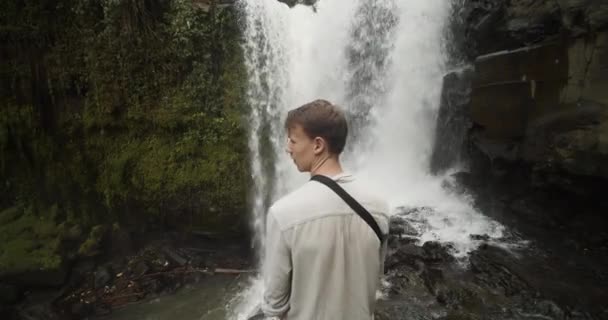 Foto jarak dekat seorang pria kulit putih muda dengan rambut coklat berdiri di tepi tebing di bawah air terjun besar di hutan Bali, Indonesia — Stok Video