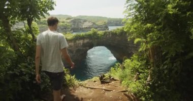 Bali Endonezya 'daki Kırık Plaj Pasih Uug' un ünlü kemeri ve okyanus kayalıklarının yakınındaki uçurumun kenarına doğru yürüyen bir erkek turistin statik görüntüsü.