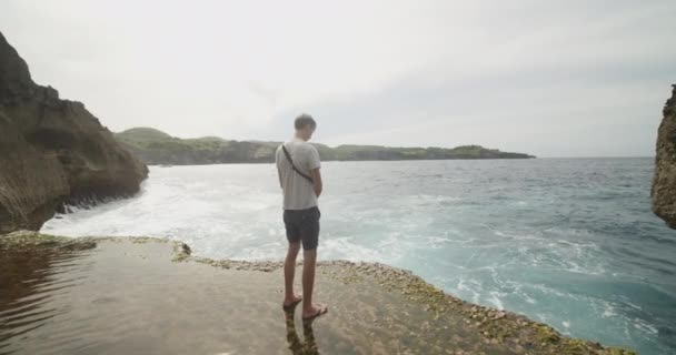 Vista posteriore di un turista bianco in piedi sulla spiaggia di scogliera allagata e guardando in un oceano a Broken Beach Pasih Uug a Bali, Indonesia — Video Stock