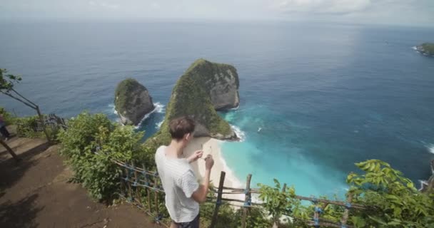 Turista masculino vestindo óculos de sol e observando a bela península tropical da baía de T-Rex na praia Kelingking em Bali, Indonésia Impressionante praia tropical vista do penhasco acima — Vídeo de Stock