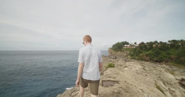 Close-up volgend schot van een man met gember haar lopend op de klif boven de oceaan langs het beroemde Broken Beach in Bali, Indonesië — Stockvideo