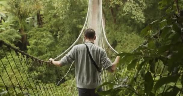 Indietro primo piano vista di un uomo che si fa strada attraverso un ponte instabile giungla di corda sotto la pioggia Giovane uomo che attraversa un ponte sospeso a Bali, Indonesia — Video Stock