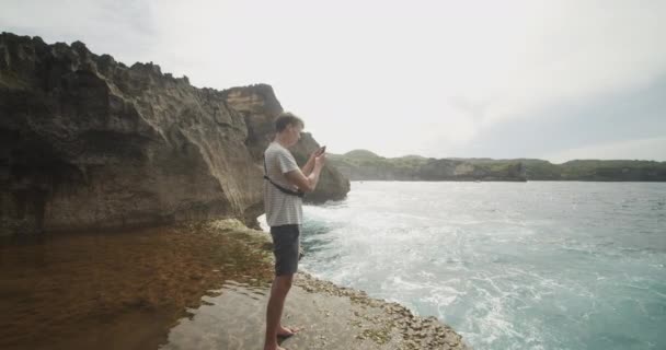 Circling handheld view of a male tourist at Broken Beach Pasih Uug in Bali, taking pictures of the view with his phone — Stock Video