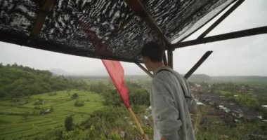 Endonezya, Bali 'deki küçük bir köye bakan, yağmurda yaban hayatı gözlem kulübesinde beyaz bir erkeğin el kamerasıyla çekilmiş görüntüsü.