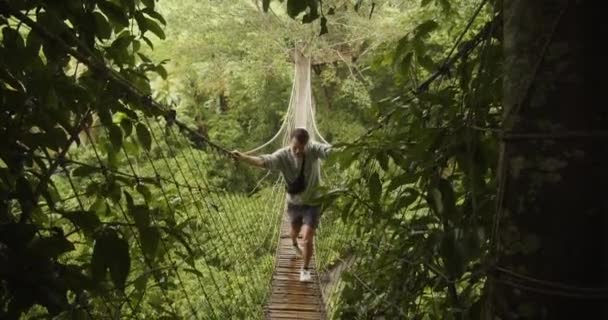 Vista estática frontal de um homem caminhando através de uma ponte instável da selva da corda na chuva Jovem pendurado nos lados de uma ponte instável — Vídeo de Stock