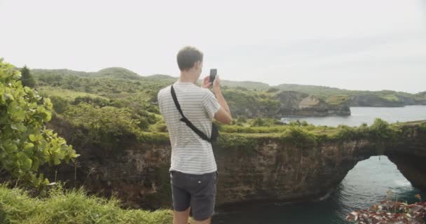 Medium close-up tiro de um turista masculino tirar fotos da praia quebrada Pasih Uug em Bali com seu telefone — Vídeo de Stock
