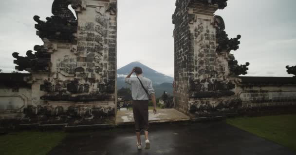 Dolly disparó después de un turista blanco caminando hacia unas famosas Puertas del Cielo en el templo Pura Penataran Agung Lempuyang en Bali con la revelación del volcán Monte Agung y los terrenos del templo — Vídeos de Stock