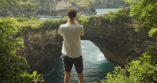 Voltar tiro de um turista masculino tirar fotos com seu telefone Homem fotografando arco e penhascos oceânicos de Broken Beach Pasih Uug em Bali Indonésia — Vídeo de Stock