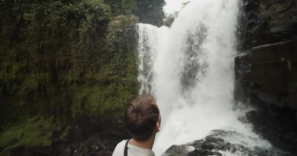 Close up shot circling above and around a young explorer pointing at the tropical waterfall and messing up his hair — Stok Video