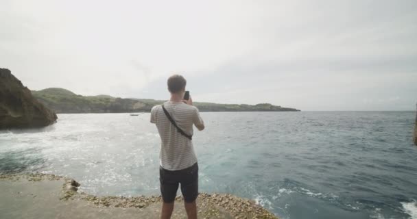 Dolly tiro médio de um turista masculino tirar fotos com seu telefone da vista em Broken Beach Pasih Uug em Bali, Indonésia — Vídeo de Stock