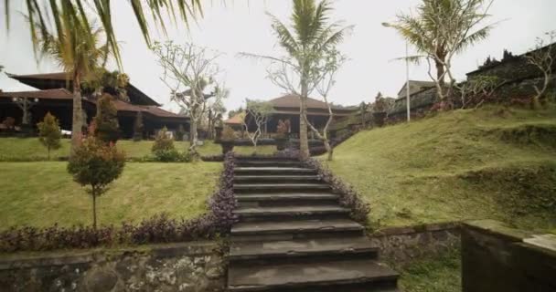 Synvinkel skott av promenader uppför trapporna i en gammal svart sten Besakih Temple i Bali Vackra tempel grunder med antika träd och blomstrande blommor — Stockvideo