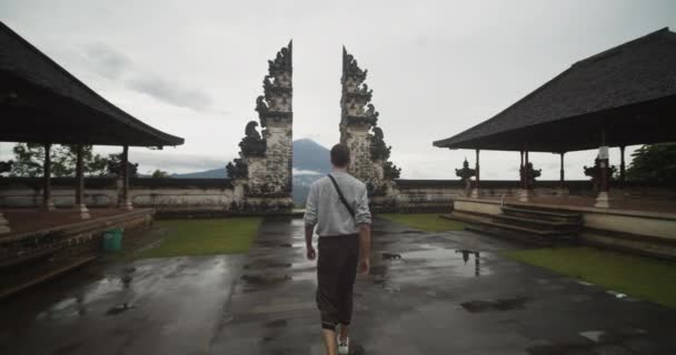Dolly disparó después de que un turista blanco caminara hacia un famoso pilar de piedra negra en el templo Pura Penataran Agung Lempuyang en Bali con el volcán Mount Agung al fondo — Vídeos de Stock