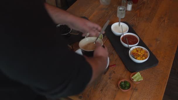 Hombre mezclando ingredientes tradicionales de la cocina mexicana en un tazón. Cuencos de salsa, arroz, frijoles negros y verduras frescas sobre la mesa. Cocine vertiendo arroz y frijoles negros en el tazón — Vídeos de Stock