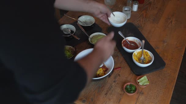 Cozinhe criando um prato tradicional mexicano em uma tigela. Homem adicionando molhos, ervas e legumes frescos em uma tigela na mesa — Vídeo de Stock