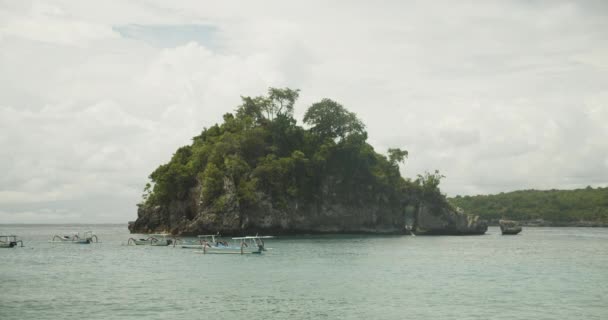 Pequeños barcos turísticos estacionados junto a una gran isla rocosa en el mar tropical. Barcos con estabilizadores en hermoso océano azul por un gran acantilado rocoso con vegetación tropical en Indonesia — Vídeos de Stock