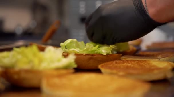 Trabajador de comida rápida en guantes añadiendo ensalada a los bollos de hamburguesa de pan. Vista de cerca de la preparación de hamburguesas en la cocina de comida rápida — Vídeo de stock