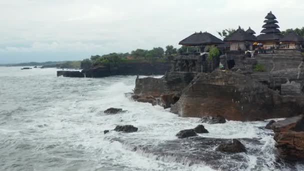 Low aerial view of strong ocean waves crashing into dark rocky cliff with Tanah Lot temple in Bali, Indonesia. Famous tourist destination in a bad weather — Stock Video