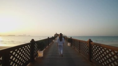 Gün batımında arka planda Dubai silueti olan tahta iskelede yürüyen bir kadın. İskelede denize doğru yürüyen uzun saçlı çekici bir kadın.