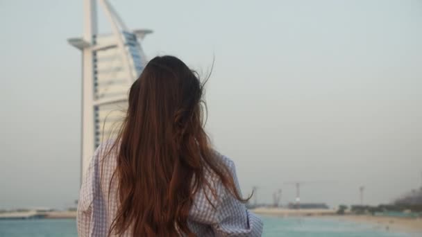 Γυναίκα δείχνει το ξενοδοχείο Burj Al Arab στο Ντουμπάι κατά τη διάρκεια του ηλιοβασιλέματος. Πίσω όψη του τουρίστα με μακριά καστανά μαλλιά κοιτάζοντας διάσημο τουριστικό αξιοθέατο στο Ντουμπάι — Αρχείο Βίντεο