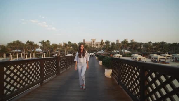 Widok z przodu kobiety spacerującej po drewnianym molo w luksusowym kurorcie turystycznym w Dubaju. Atrakcyjna kobieta z długimi brązowymi włosami spacerująca w luksusowym kurorcie turystycznym w Emiratach Arabskich — Wideo stockowe