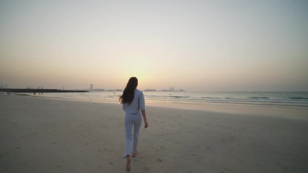 Gün batımında Dubai 'de sahilde koşan bir kadın. Sahilde okyanusa doğru koşan zarif elbiseli çekici bir esmer. — Stok video
