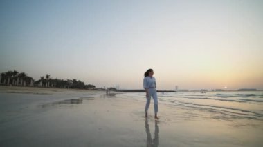 Gün batımında plajda koşan zarif elbiseli bir kadının ön görüntüsü. Sahilde koşan zarif elbiseli çekici bir esmer. Arka planda Dubai silueti var.