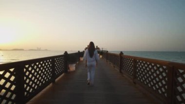 Ahşap iskelede yürüyen ve Dubai 'deki Burj Al Arap oteline bakan bir kadın. Dubai 'deki kumsalın kıyısındaki ahşap iskelede yürüyen kadın turistin arka görüntüsü.