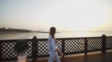Gün batımında Dubai 'de okyanus iskelesinde yürüyen bir kadın. Burj Al Arap oteline bakan bir kadın ve tahta bir iskeleden kumsala bakan bir kadın.