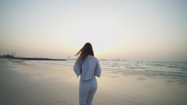 Kumsalda koşan ve kameraya bakan bir kadın. Gün batımında Dubai 'de plajda koşan zarif elbiseli çekici esmer kadın.