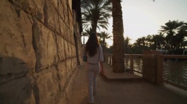 Dubai 'de lüks bir mahallede nehir kenarında yürüyen bir kadın. Sıcak günbatımında nehir boyunca yürüyen kadının arka görüntüsü