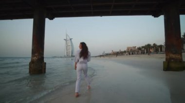 Dubai 'de kumsalda Burj Al Arap otelinin önünde yürüyen çekici genç bir kadın.